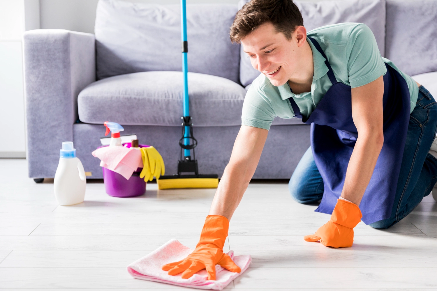 Simply Clean Niederrhein - Professionelle Reinigung - Ein lächelnder junger Mann in grünem Poloshirt, blauer Schürze und orangefarbenen Gummihandschuhen kniet auf einem hellen Boden und wischt ihn mit einem rosa Tuch. Im Hintergrund sind ein graues Sofa, ein blauer Wischmopp und verschiedene Putzutensilien in einem violetten Eimer zu sehen.