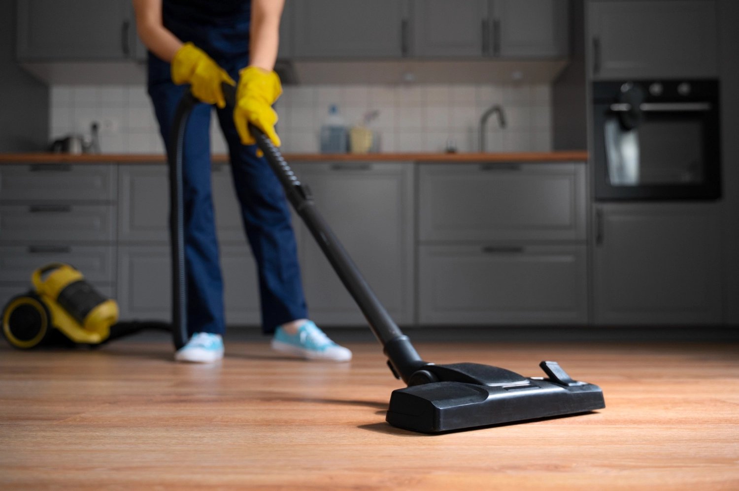 Simply Clean Niederrhein - Professionelle Reinigung - Eine Person in blauer Kleidung und gelben Handschuhen saugt einen Holzboden in einer Küche mit grauen Schränken.
