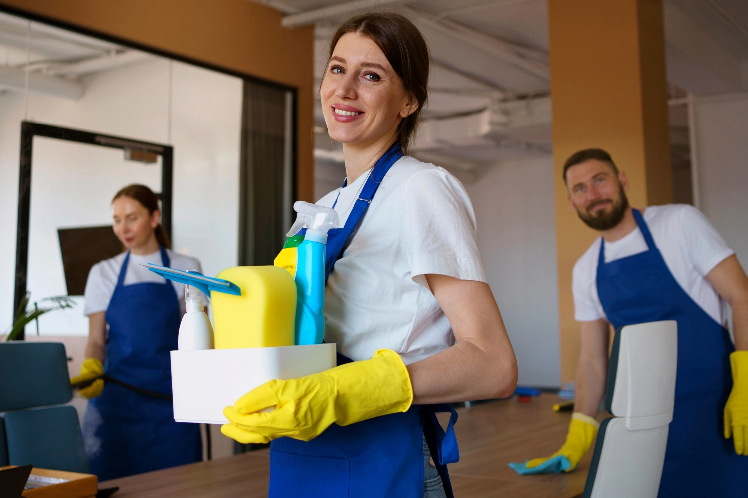 Simply Clean Niederrhein - Professionelle Reinigung - Eine lächelnde, professionelle Reinigungskraft in blauer Schürze und gelben Handschuhen hält in einem hellen Büro einen Behälter mit Reinigungsmitteln, während zwei Kollegen im Hintergrund arbeiten.