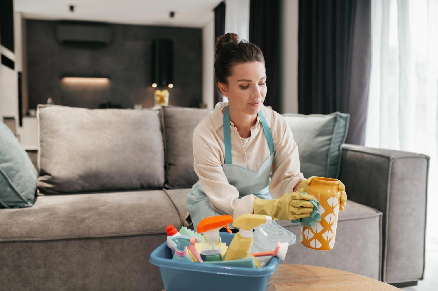 Simply Clean Niederrhein - Professionelle Reinigung - Eine Frau in Schürze und Handschuhen reinigt eine gelb gemusterte Vase in einem Wohnzimmer; daneben steht ein Korb mit Putzutensilien.