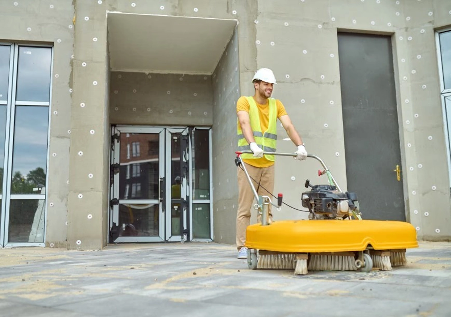 Simply Clean Niederrhein - Professionelle Reinigung - Ein Bauarbeiter mit Schutzhelm und Sicherheitsweste bedient eine gelbe, handgeführte Kehrmaschine, um einen gepflasterten Gehweg vor einem modernen Gebäude zu reinigen.