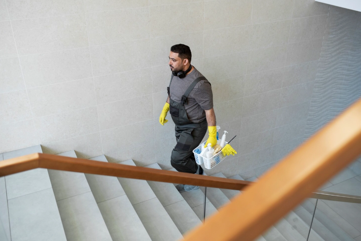 Simply Clean Niederrhein - Professionelle Reinigung - Ein professioneller Reinigungsmann in grauem Overall und gelben Handschuhen geht eine moderne Treppe hinauf und trägt dabei einen weißen Wagen voller Reinigungsmittel.