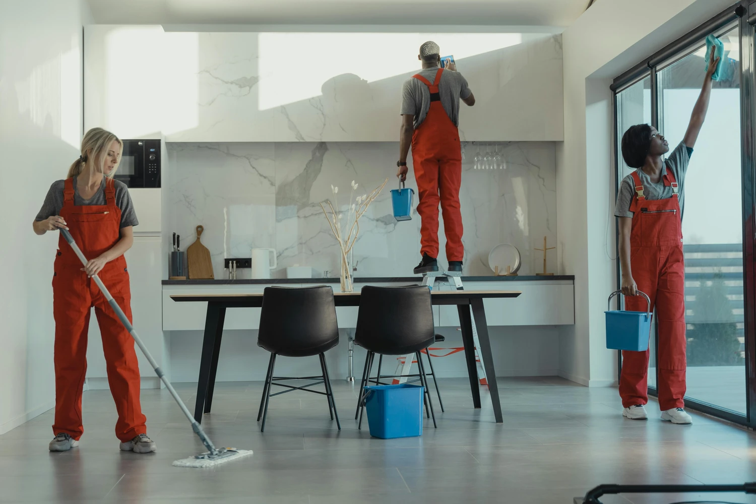 Simply Clean Niederrhein - Professionelle Reinigung - Ein Team aus drei professionellen Reinigungskräften in roten Overalls arbeitet zusammen, um eine moderne, hochwertige Küche und einen Essbereich zu reinigen.